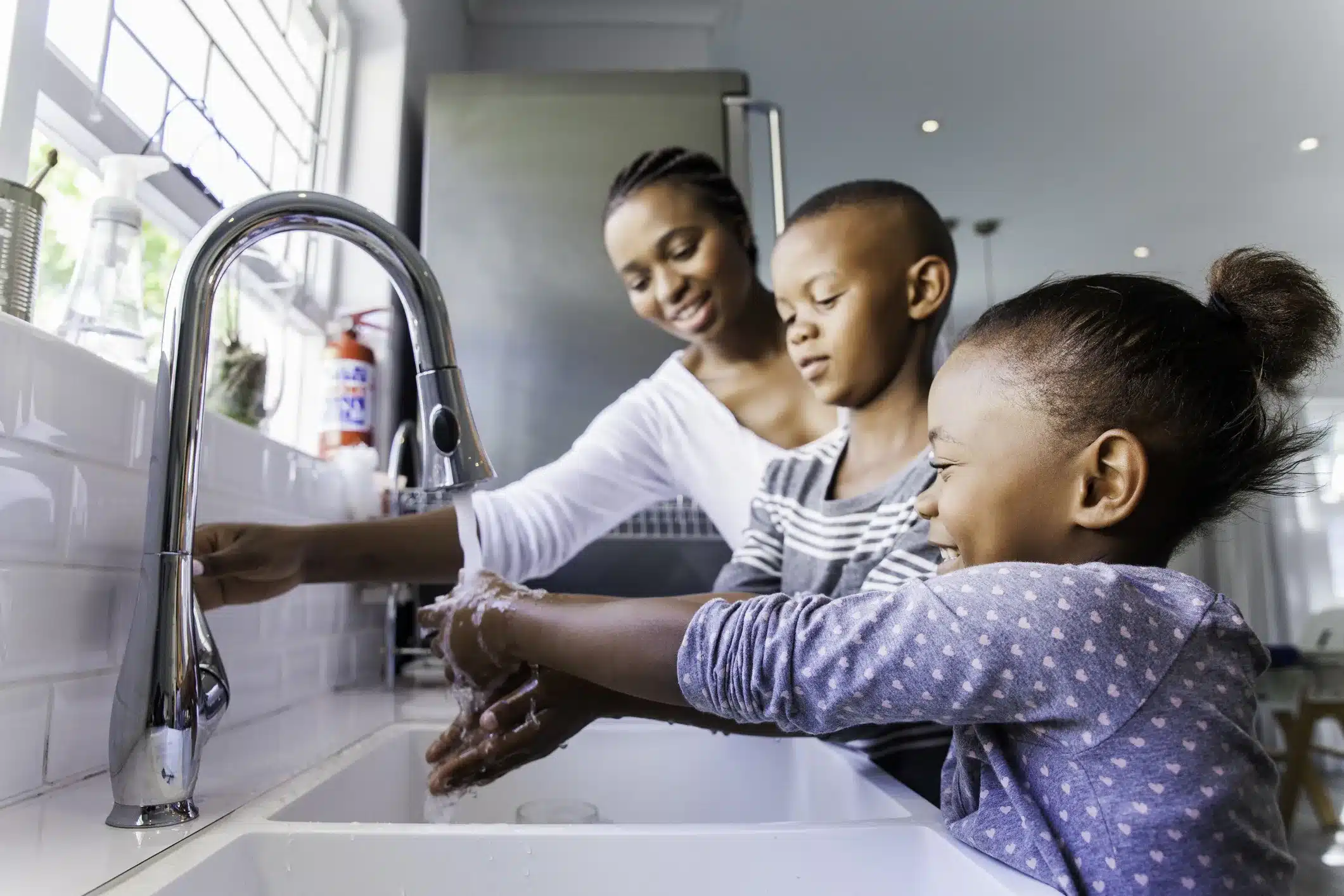 A mom helps her son and daughter wash their hands at the kitchen sink.